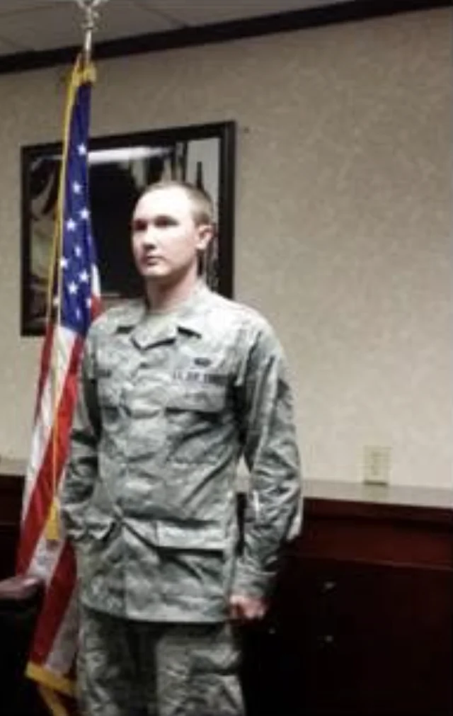 A young Josh Poulin in Air Force uniform, standing at attention beside the United States flag